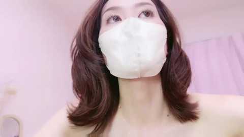 Media: Video of a young Asian woman with shoulder-length brown hair, wearing a white face mask, in a softly lit, pastel-colored room with a light pink curtain in the background.