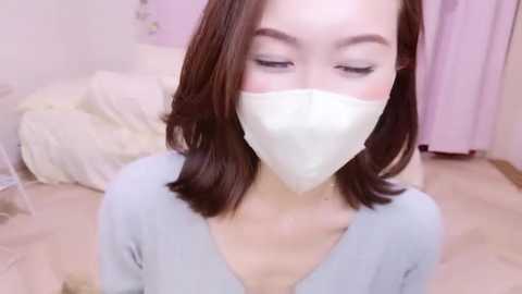 Media: Video of an Asian woman with shoulder-length dark hair, light skin, and closed eyes, wearing a white face mask and light blue top. Background features a soft, pastel-pink wall and a bed with white sheets.