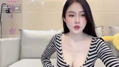 Media: A video of an East Asian woman with long black hair, wearing a black and white striped top, posing in a modern living room with beige walls.