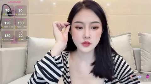 Media: A video of an Asian woman with long black hair, fair skin, and full makeup, adjusting her hair on a white couch.