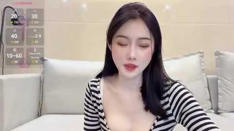 Media: A video of an East Asian woman with long black hair and fair skin, wearing a black and white striped cardigan, posing in a modern living room with white furniture.