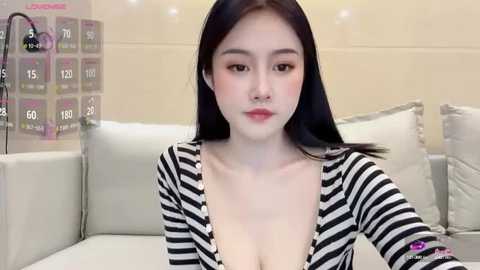 Media: Video of a young Asian woman with pale skin, long black hair, and light makeup, wearing a black and white striped top, sitting on a white couch in a modern living room.