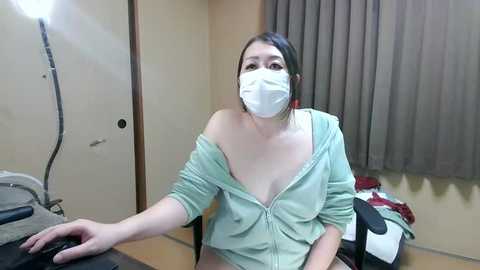 Media: Video of an Asian woman with a light skin tone, wearing a white mask, off-shoulder mint green top, sitting at a desk in an office setting, with a laptop and gray curtains in the background.