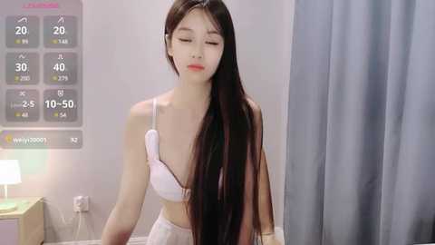 Media: Video of an East Asian woman with long black hair, fair skin, and a slim physique, wearing a white bra and panties. She stands in a minimalist bedroom with a digital display showing temperature and humidity.