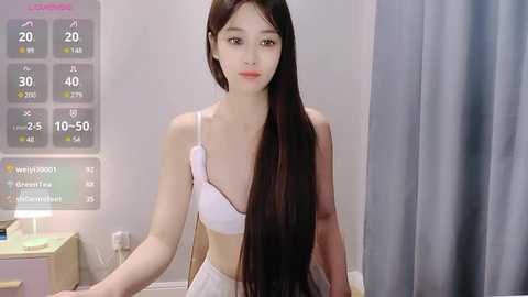 Media: A video of a young East Asian woman with long, straight black hair, wearing a white bra and panties, standing indoors, with a weather display and gray curtain in the background.