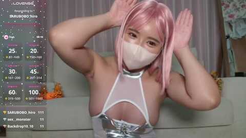 Media: A video of an Asian woman with pink hair, wearing a white face mask, a silver halter top, and a white skirt, posing playfully indoors.