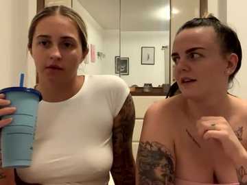 Media: Video of two women in a bathroom; one with a white top, the other topless, both holding blue drinks, discussing tattoos.