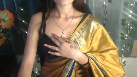 Media: A video of a woman with light brown skin, wearing a gold sari with black blouse, silver necklace, and a hand on her chest, indoors with floral decorations and fairy lights in the background.