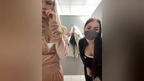 Media: A video of two women in a bathroom; one wearing a pink dress, the other in a black top, both with masks on. Background shows hanging clothes.