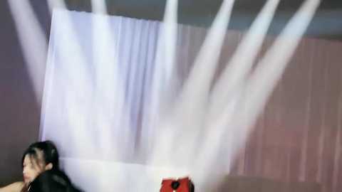 Media: Video of a performer with long black hair, wearing a black top, being sprayed by white mist from a stage prop, against a white backdrop.