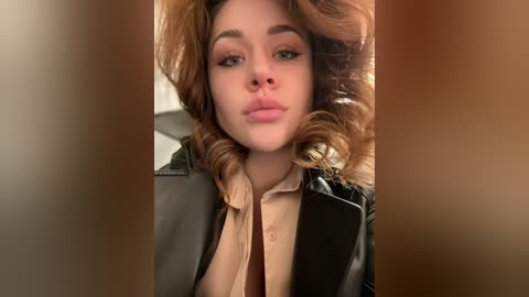 Media: A close-up video of a fair-skinned woman with voluminous, wavy, auburn hair, wearing a black leather jacket over a beige shirt. She has a neutral expression and is slightly out of focus.