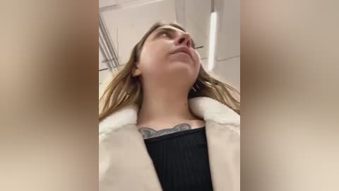 Media: Video of a young Caucasian woman with light skin, brown hair, and a tattoo on her chest, wearing a black top and white fur coat, looking upward.