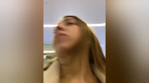 Media: A blurry video of a woman with long brown hair, wearing a beige jacket, captured from a low angle, possibly in a public space with a bright, modern ceiling.