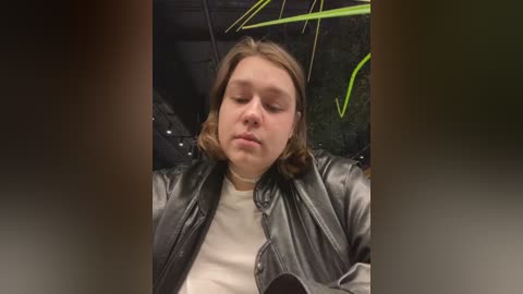 Media: Video of a young Caucasian woman with shoulder-length brown hair, wearing a black leather jacket over a white t-shirt, lying on a black leather couch, with green plant leaves visible in the background.