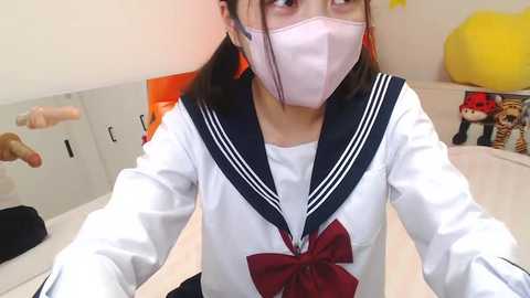 Media: Video of an East Asian woman in a Japanese schoolgirl cosplay outfit, with a pink mask, white blouse, red bow, and blue sailor collar. Background includes a white locker and plush toys.