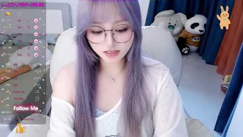 Media: Video of a young Asian woman with long, lavender hair, wearing glasses, a white off-shoulder top, sitting in a white chair, surrounded by plush toys in a room with blue curtains.