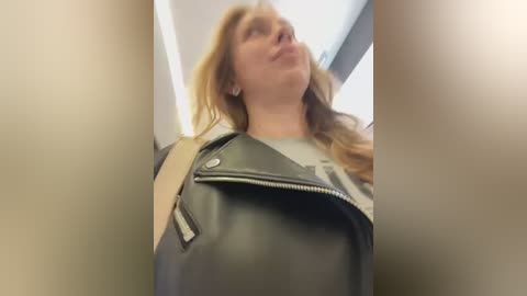 Media: Video of a Caucasian woman with long, wavy red hair, wearing a black leather jacket over a white shirt, captured from a low angle, creating a distorted perspective.