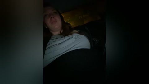 Media: A dimly lit video of a young woman with long brown hair, wearing a white T-shirt and black jacket, lying on a bed with a dark wooden headboard.