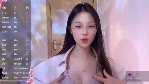 Media: A video of an East Asian woman with long black hair and fair skin, wearing a light pink bra and open white shirt, standing in a mirror-lit room with a blurred background.