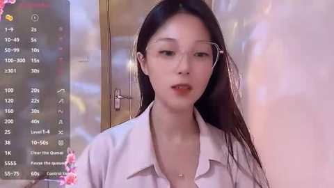 Media: Video of a young East Asian woman with long black hair and glasses, wearing a white shirt, captured indoors with a blurred background.