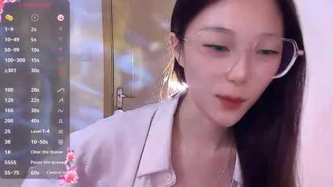 Media: A video of an Asian woman with fair skin, long black hair, wearing glasses and a white blouse, looking slightly downcast, in a blurred indoor setting.