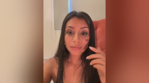 Media: Video of a Latina woman with long black hair, fair skin, and dark eyes, wearing minimal makeup, sitting in a red chair, touching her face, with blurred background.