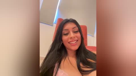 Media: Video of a smiling Latina woman with long black hair, wearing a pink bra, sitting in a red chair in a brightly lit room with white walls and windows.