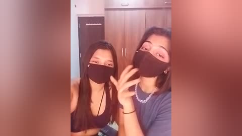 Media: A video captures two young women with long black hair, wearing black masks, smiling and touching noses in a playful manner. They are in a room with wooden cabinets and a dark door.