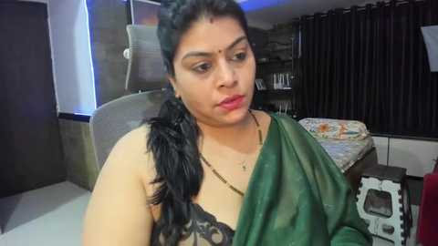 Media: Video of a South Asian woman with medium skin tone, wearing a green saree, seated on a beige chair, in a modern living room with dark curtains, a floral bedspread, and a TV.