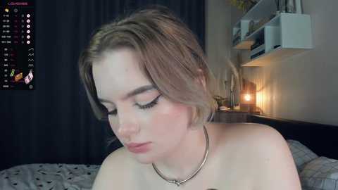 Media: Video of a young woman with fair skin, light brown hair, and closed eyes, wearing a thin silver necklace, in a dimly-lit bedroom with dark curtains, a bed with a patterned sheet, and a digital clock on the wall.