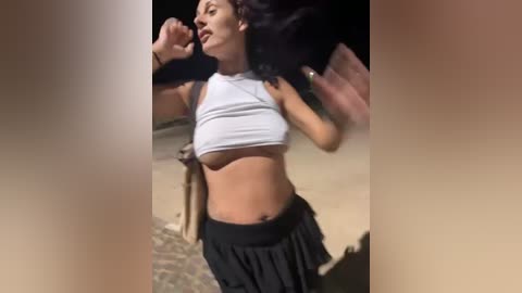 Media: Video of a young woman with light brown skin, dark hair, wearing a white crop top and black skirt, dancing outdoors at night.