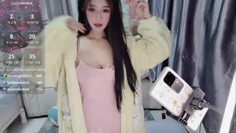 Media: A video of a young East Asian woman with long black hair, wearing a pink spaghetti strap dress and a light yellow fur coat, posing in front of a gray curtain, with a phone camera visible.