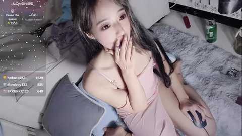 Media: A video of an Asian woman with long hair, wearing a pink tank top, kneeling on a plush gray rug, covering her mouth in a playful manner.