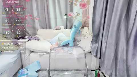 Media: Video of a messy, white, plush sofa with a blue, plush dragon toy, surrounded by scattered toys and clothing, in a room with gray curtains and pink flowers.