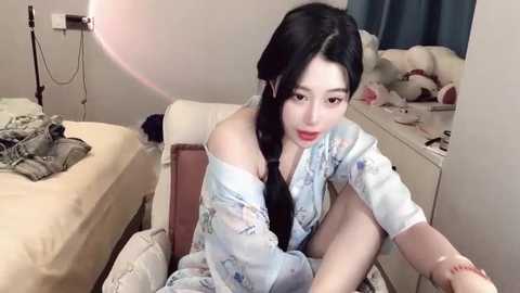 Media: A video of an East Asian woman with long black hair, fair skin, and a slender physique, sitting on a bed in a cozy room, wearing a light floral robe, surrounded by plush toys and a cluttered dresser.