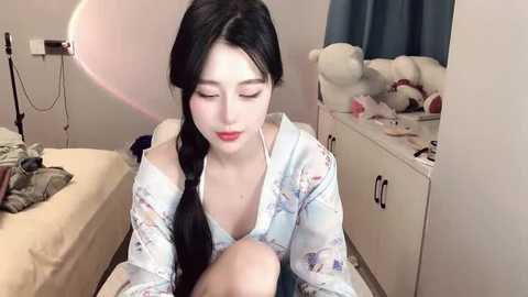 Media: A video of an East Asian woman with long black hair in a floral pajama top, sitting on a bed in a cozy, dimly lit room with stuffed animals and a nightstand.