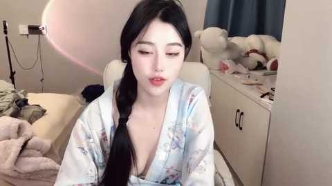 Media: Video of an East Asian woman with pale skin, long black hair, and a V-neck robe, sitting in a messy bedroom with stuffed animals, a bed, and a medical device in the background.