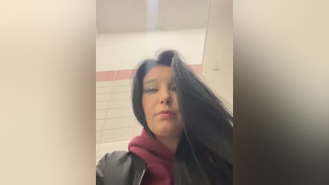 Media: A video of a young woman with long black hair, wearing a black jacket over a maroon hoodie, standing in a tiled restroom with white and pink tiles. Her face is slightly obscured by the camera angle.