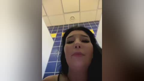 Media: A video of a young Asian woman with long black hair, wearing a black tank top, standing in a bathroom with blue-tiled walls and a white ceiling. She has a neutral expression.