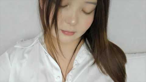 Media: A video of an Asian woman with long, straight brown hair and fair skin, wearing a white button-up shirt. She has her eyes closed, appearing relaxed. The background is plain white, focusing attention on her.