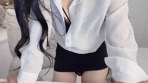 Media: Video of a woman in a white button-down shirt, black shorts, and black panties, with long black hair, leaning over a white couch in a modern, minimalistic living room.