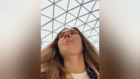 Media: Video of a young woman with long brown hair, wearing a white t-shirt and plaid shirt, looking up, surrounded by a geometric glass ceiling.