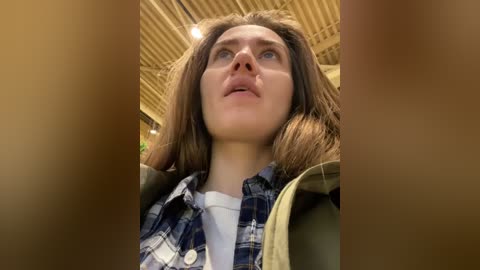 Media: Video of a young woman with light brown hair, wearing a blue plaid shirt, looking up thoughtfully under a wooden ceiling.