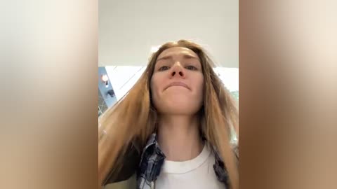 Media: A video of a young Caucasian woman with long, straight blonde hair, wearing a white t-shirt and a denim jacket, with a neutral expression, taken indoors with blurred background.