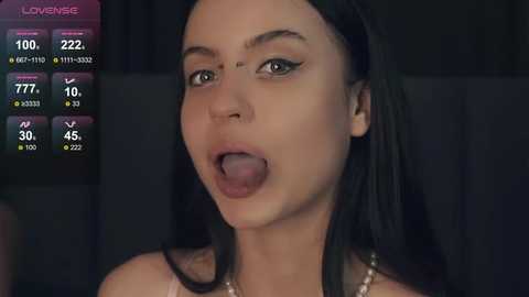 Media: Video of a young woman with long black hair, brown eyes, and dark makeup, sticking out her tongue. Background shows a dark room with a TV screen displaying \"Loverense.\