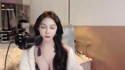 Media: Video of a fair-skinned, East Asian woman with long black hair, wearing a white cardigan over a low-cut top, recording herself with a camera on a tripod in a minimalistic, beige room.