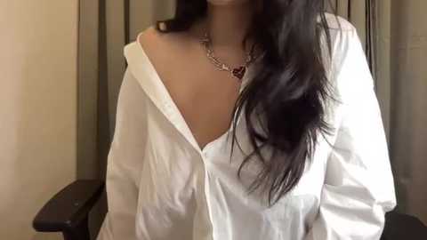 Media: Video of a woman with long, straight black hair, wearing a white, unbuttoned shirt revealing her cleavage, and a silver necklace with a heart pendant, standing indoors.