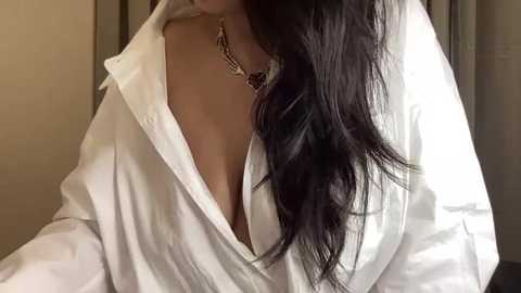 Media: Video of a woman with long, wavy black hair, wearing a loose white shirt that reveals cleavage, against a neutral background.