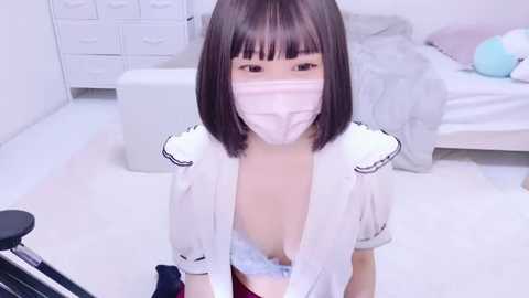 Media: Video of an Asian woman with a mask, short black bob, and a white top, exposing cleavage, in a minimalist white room with a bed and drawers.