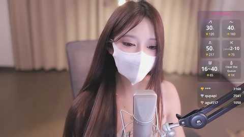 Media: A video of a young Asian woman with long, straight brown hair, wearing a white mask, seated at a desk, singing into a microphone, with a digital thermometer display on screen.